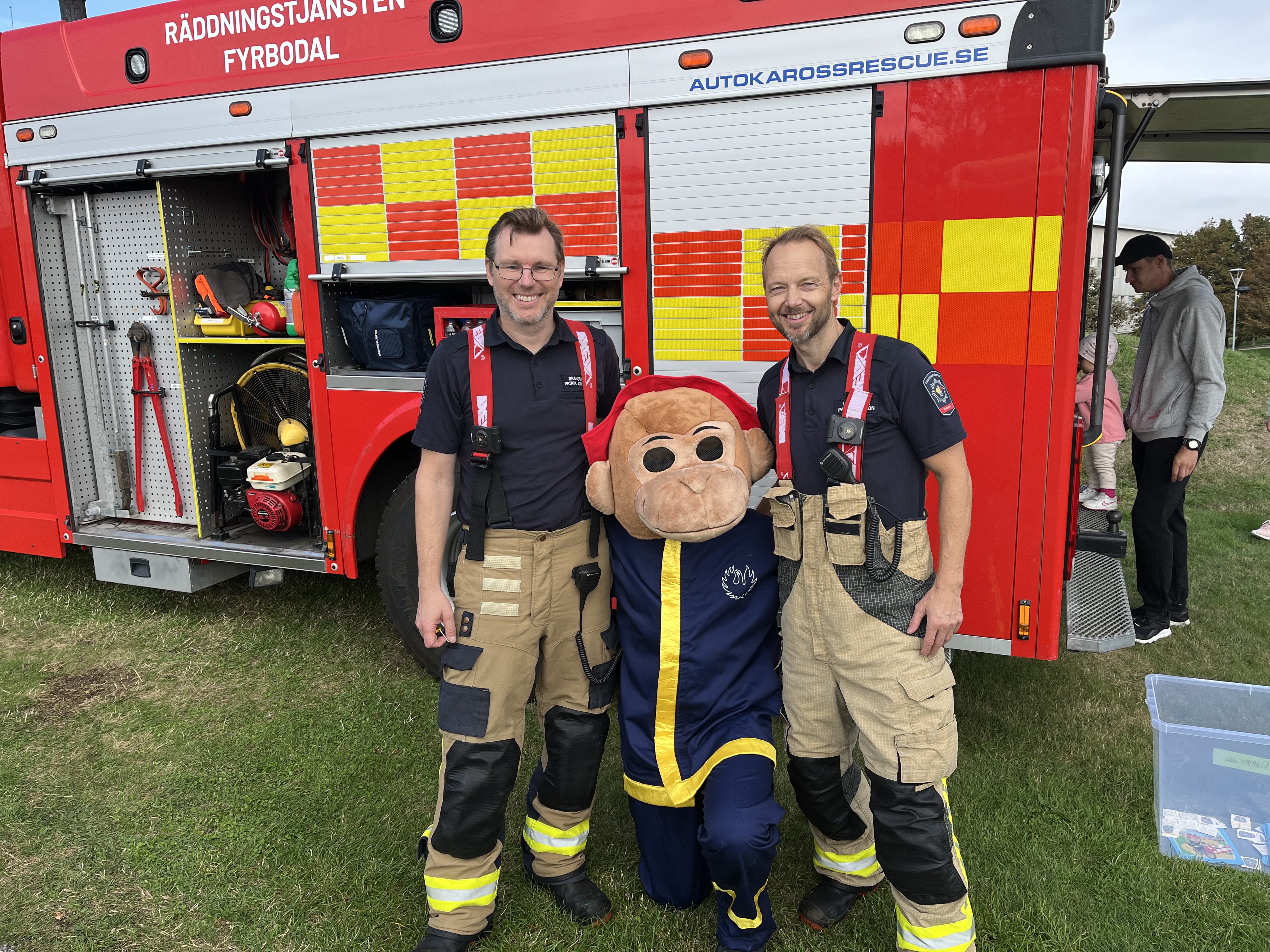 Två brand står framför brandbilen med en maskot emellan sig.