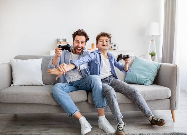 En pappa och son sitter i en grå soffa och spelar tvspel
