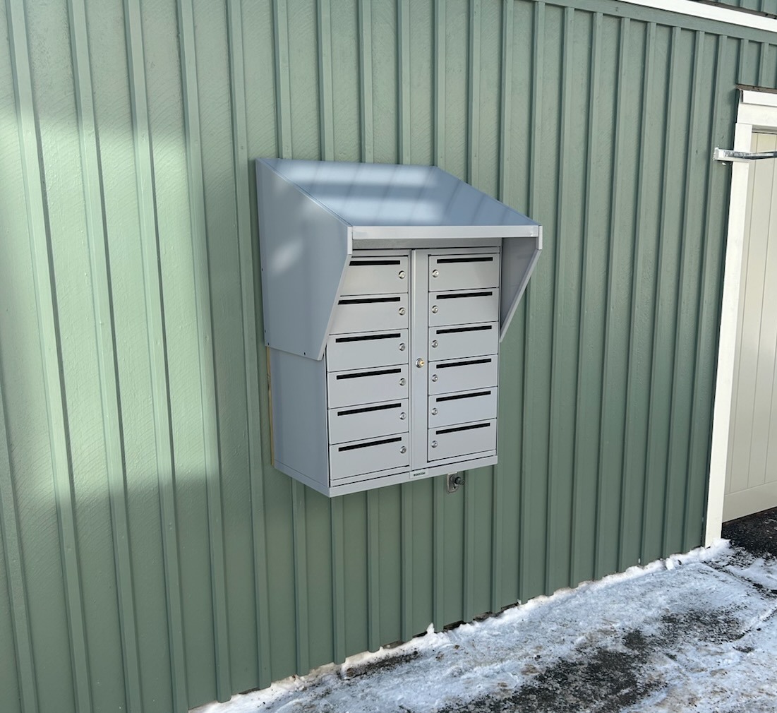 Ett aluminiumfärgat brevlådeskåp är monterat på en grönmålad träfasad.