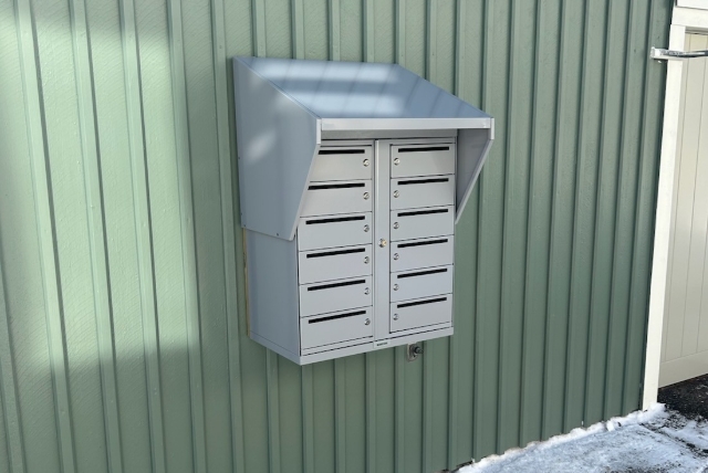 Ett aluminiumfärgat brevlådeskåp är monterat på en grönmålad träfasad.